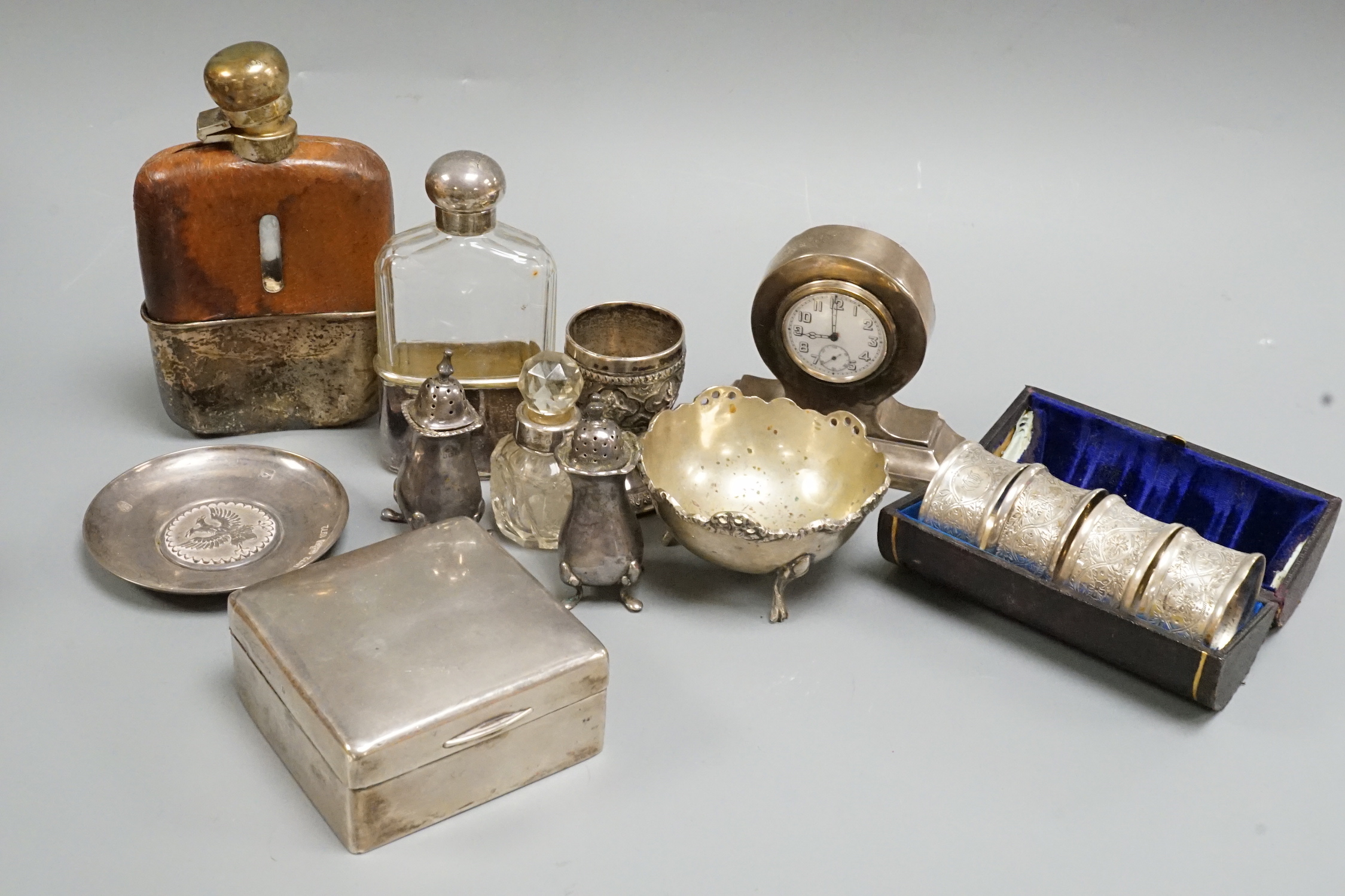 A George V silver mounted small mantel clock, height 93mm, a pair of silver pepperettes and eight other items including a silver mounted cigarette box, a cased set of four chased silver napkin rings and two plated hip fl